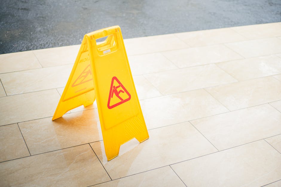 A bright yellow, portable wet floor caution sign placed on a tiled outdoor patio area. The sign features a red triangular warning symbol with a figure slipping, alerting passersby to potential slippin