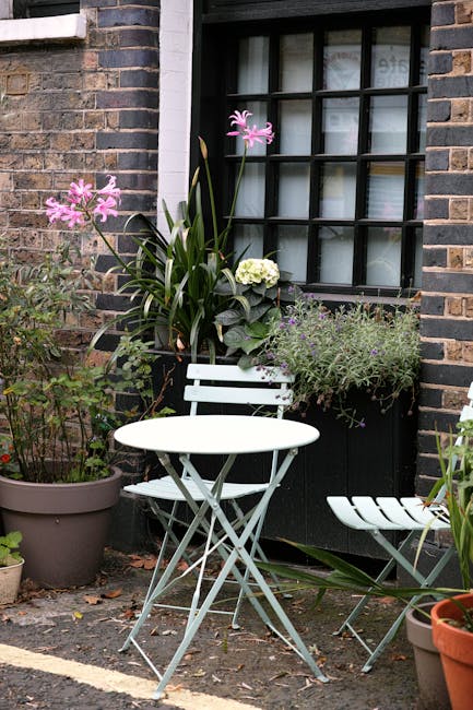 The image depicts a quaint front garden beside a brick house with a black grid-style window. A small white metal table and matching chair are positioned on the paved area, surrounded by various potted